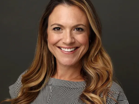 A smiling woman with shoulder-length wavy hair poses in a studio portrait against a dark background.