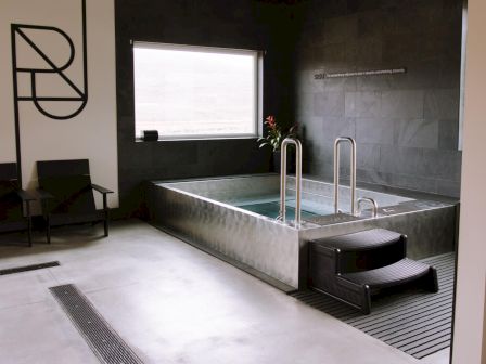 A sleek spa-like bathroom with a stainless hot tub, shower handles, tiled walls, a large window, and seating with a wall logo.