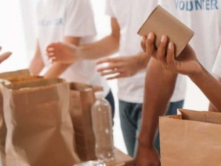 Volunteers wearing &ldquo;VOLUNTEER&rdquo; shirts are packing food supplies into paper bags and boxes at a distribution table.